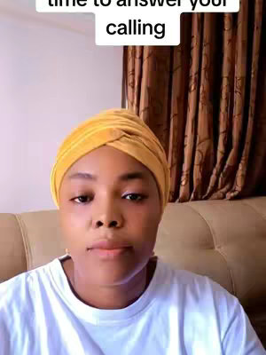 A woman with a yellow head wrap and a white t-shirt looks directly at the camera. Above her head is a white rectangle with black text. The background includes brown curtains and a beige sofa.