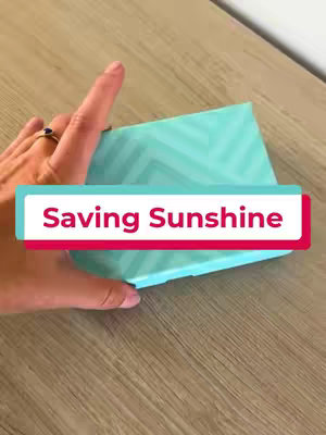 A person's hand holding a partially visible, bright turquoise box with a chevron pattern, placed on a wooden surface. A red and white banner with the text 'Saving Sunshine' covers the box, indicating a brand announcement.