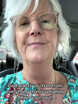 A selfie-style video features a woman with short, white hair wearing clear-framed glasses and dangling earrings. She is positioned inside what appears to be a car, with the background slightly blurred. She is wearing a teal and floral patterned top, and the text overlay at the bottom reads 'God wants you free from everything that is weighing down your soul!'