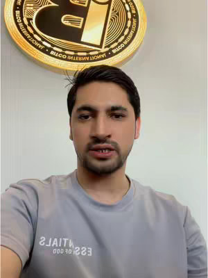 A man in a grey t-shirt speaks directly to the camera, with a circular gold Bitcoin logo visible behind him. The setting appears to be an office with a plain wall.