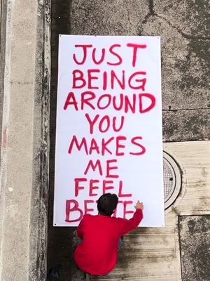 An overhead shot captures a person in a red shirt writing on a large white poster propped against a rough, gray concrete surface. The poster has handwritten text in red. A drain cover is visible. The composition is simple, with the focus on the text and the person interacting with it.