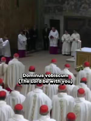 The cover image shows a group of people, likely religious figures, in a church setting. In the foreground, many individuals wear white robes and red hats. Some people are behind the red-hatted individuals wearing white. In the background, people are visible near an altar. A text overlay at the bottom reads "Dominus vobiscum (The Lord be with you)".
