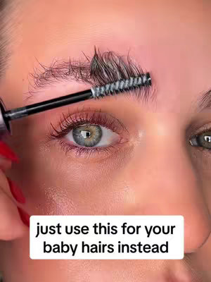 A close-up shot of a person's face with focus on the eye, eyebrow, and part of the nose. The person has long eyelashes and light green eyes. There is a black eyebrow brush being used on one eyebrow, and visible baby hairs are sticking up. On the bottom of the image, there is a white text box that reads 'just use this for your baby hairs instead'.