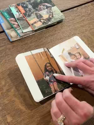 A person is holding open a photo journal, with a photo of a young girl on a swing visible. Other photo journals are stacked to the side. The person's hands, with a ring, are visible as she points at a photo. The background contains a wooden surface.