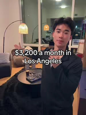 A man stands in a well-lit, modern apartment, gesturing with one hand. The background includes a table with items, a large window overlooking a city skyline, lamps, and soft seating.