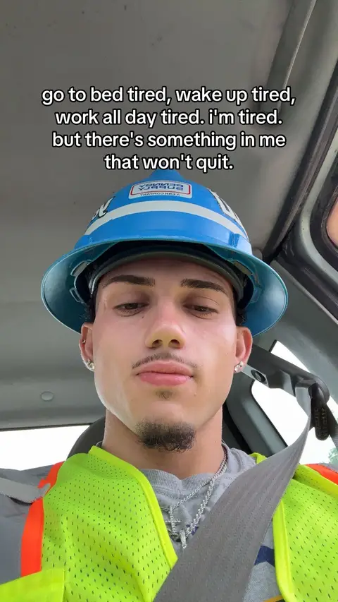 A selfie of a young man inside a vehicle, likely a truck or car. He's wearing a blue hard hat and neon green safety vest. The man has a serious expression, and text is overlaid at the top.