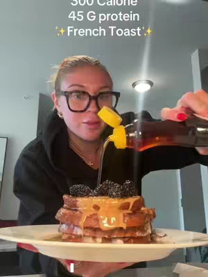 A woman with blonde hair and glasses is shown, pouring syrup over a stack of French toast that is topped with berries. The toast sits on a white plate. The text overlay at the top states the calorie and protein value of the meal.