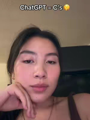 A face close-up of a young woman with dark hair, positioned slightly off-center. She has fair skin, dark eyes, and a subtle expression. The upper portion of the frame includes text that states "ChatGPT = C's" followed by an emoji. The lighting is soft, highlighting her features. The background is out of focus, suggesting an interior setting.