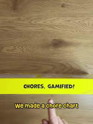 The video shows a wooden floor with a yellow banner that reads "CHORES, GAMIFIED!". Below the banner, the text "We made a chore chart" is present, with a finger pointing to the banner.