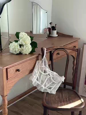 A well-lit interior scene featuring a wooden vanity with a tri-fold mirror. A bouquet of white flowers sits on the vanity, and makeup brushes are visible. A white lace lingerie piece hangs on a wooden chair in front of the vanity. The overall aesthetic is soft and feminine.