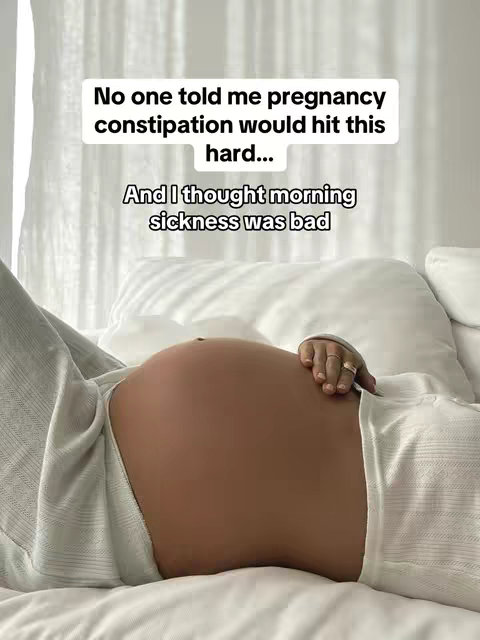 A pregnant woman lies in bed, her bare belly prominent, the focus of the image. She's resting on white bedding, hands casually placed, rings visible, providing a sense of intimacy and personal space.