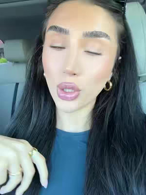 A close-up shot of a woman inside a car, looking towards the camera with closed eyes and slightly parted lips. She has dark hair, thick eyebrows, and is wearing gold hoop earrings. The background is out of focus, showing the interior of the car.