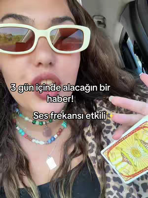 A selfie of a woman inside a car, primarily focusing on her face and upper body. She wears light-colored, thick-framed sunglasses and a layered necklace. Text overlays are added to the image. In her hand she holds a tarot card.