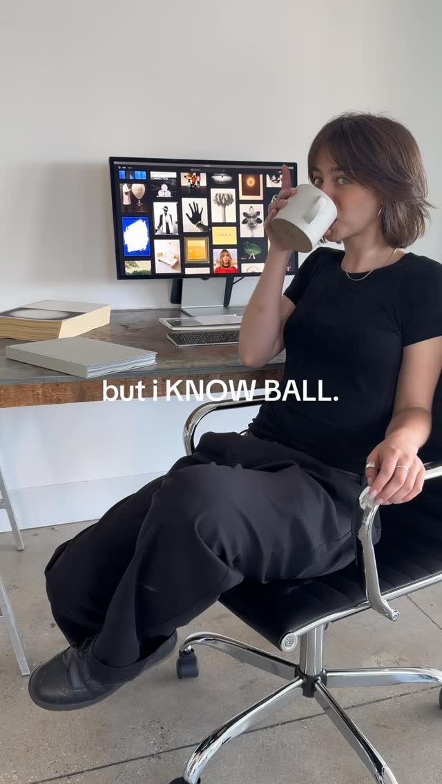A young woman is seated in a black office chair, holding a white mug and taking a sip. She is wearing a black t-shirt and black pants. Her right hand rests on the chair's arm, and she has rings on her fingers. Behind her is a wooden desk with a computer monitor displaying a mood board. The monitor has an array of images on its screen. There are books to the side with a white wall in the background lit by natural light. The scene conveys a casual, stylish office setting.