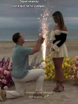 A man kneels to propose to a woman on a beach at sunset. Fireworks are going off behind them. The woman is looking down at the man.