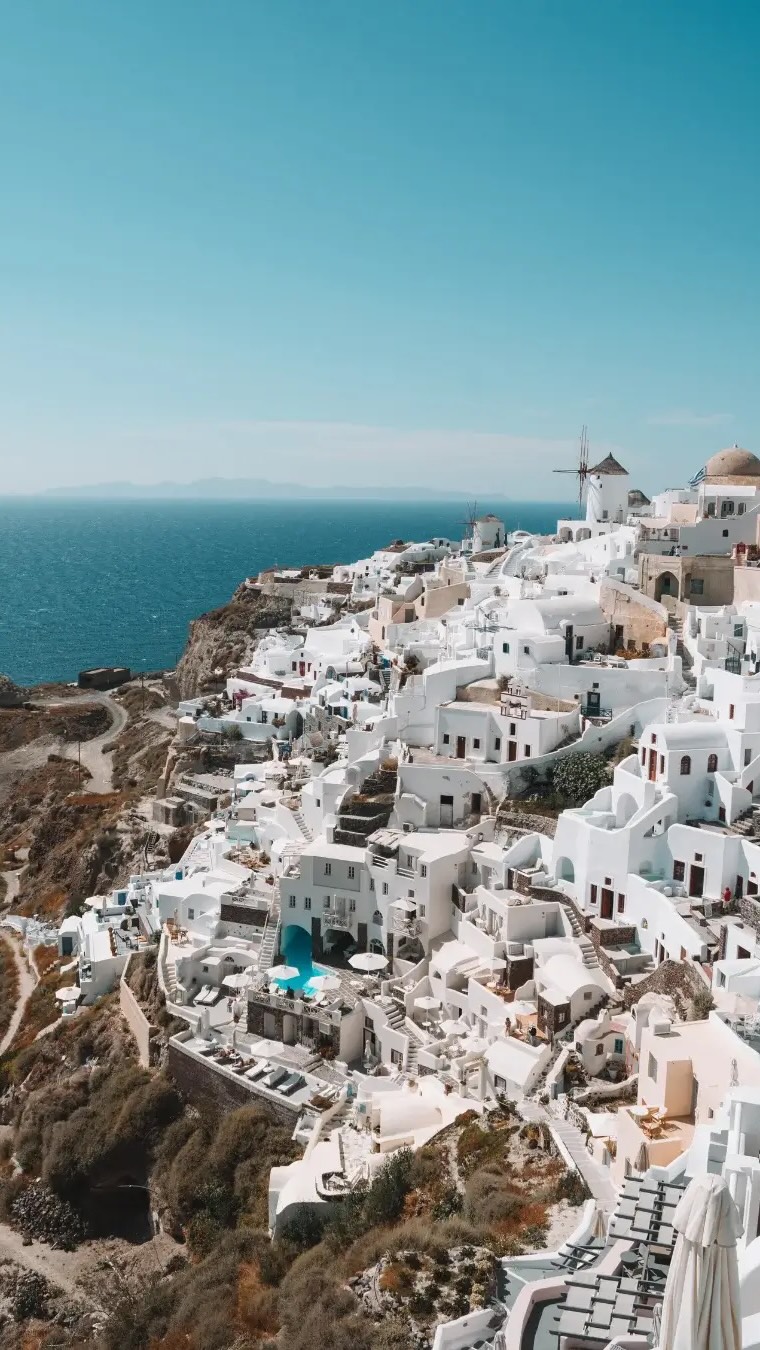 A high-angle shot captures the iconic white buildings of Santorini, Greece, cascading down a hillside towards the deep blue Aegean Sea. The buildings are closely packed, featuring traditional whitewashed walls, blue-domed roofs and arches. In the distance, the sea meets a clear, light-blue sky. The composition is simple and elegant, with the natural colors of the region dominating the scene.