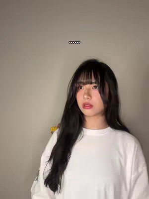 A young woman with long, dark hair and bangs is looking slightly off-camera. She is wearing a white long-sleeved shirt against a neutral background. Above her head are a series of dots.