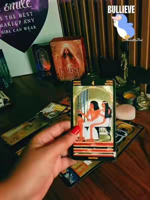 A hand with red painted nails holds a tarot card showing an illustration of two figures. Other cards are spread out on a wooden surface, with objects like a candle and a box with a picture of a woman in the background. The image is lit with warm lighting.