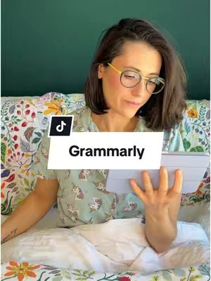 A woman with short, dark hair wearing glasses sits on a bed. She's in a light green shirt with a pattern of small white unicorns and is holding a tablet. The background includes green wall, sheets with floral pattern. A TikTok logo with the text Grammarly, is added on the cover image.