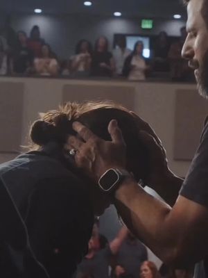 A photo from a low angle shows a man placing his hands on another person's head, possibly in a church setting. The person's head is bowed, and the hands are resting directly upon it. In the background, a blurred image of an audience, possibly in a church or large meeting space.