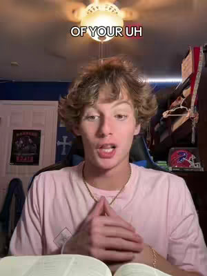 A young man with curly brown hair is centered in the frame, looking directly at the viewer with a slight smile. He's wearing a light pink t-shirt and a gold chain. The background includes a white door, a poster with text, and various items on shelves, and a ceiling fan is visible overhead with text overlayed across the top.