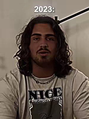 A man with long, wavy dark hair and a mustache is centered in the frame, wearing a t-shirt with a graphic design. Above him, white text states '2023:', indicating a year or timeframe. The background is blurred, suggesting an indoor setting, and a portion of a dark object is visible above his head. The man looks directly at the camera.
