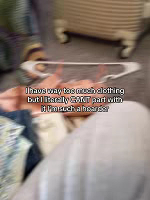 The image has a blurred, close-up perspective, with a focus on a person's hands holding an item, likely clothing, near a white hanger. The background appears out of focus but hints at a setting containing more items, possibly a room or closet. The image has a warm, slightly desaturated color scheme. The text overlayed on the image mentions 'too much clothing'.