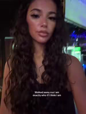 A selfie-style image of a woman with long, wavy dark hair. She's looking towards the camera with a neutral expression. The background is blurred, suggesting an indoor setting, with some indistinct lighting and possibly a bar area or some kind of venue with neon signs. Text overlayed reads 'Walked away cuz I am exactly who tf I think I am'.