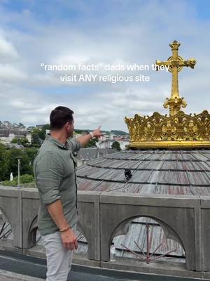 A man in a green shirt and white pants stands on a stone structure with a golden cross and large crown in the background, pointing with his right hand. The sky is overcast.