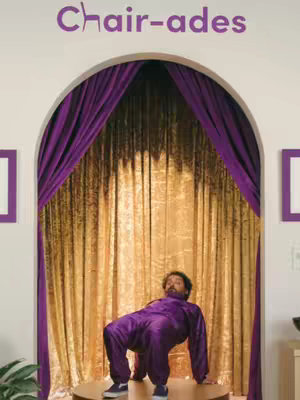 The cover image shows a man dressed in a purple jumpsuit, posed as a chair on a circular platform. He is in front of a backdrop of a large golden curtain framed by an archway. The text 'Chair-ades' is above the arch.