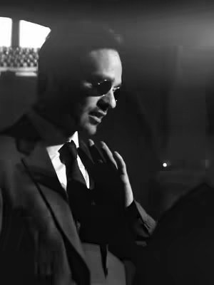A black and white image of a man in a suit and sunglasses. He is holding his hand up to his face and looking down. The lighting is dim, suggesting a dramatic or mysterious atmosphere.