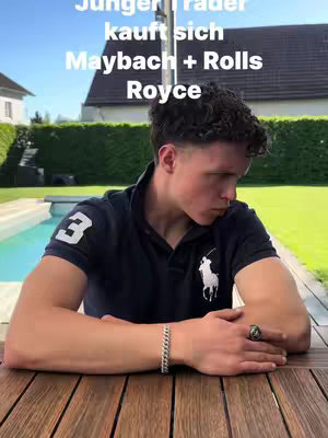 A young man with short curly hair is seated at a wooden table, wearing a dark blue polo shirt with the number 3 and a white emblem. He has a silver bracelet and a ring on his finger. The background shows a swimming pool and a well-kept lawn framed by a house.
