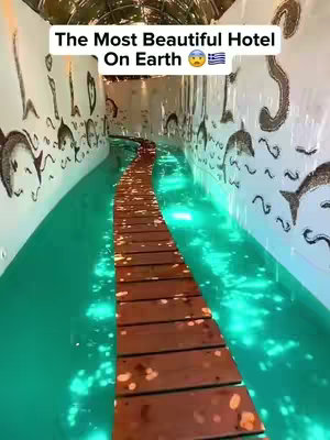 The image displays an interior view of a hotel featuring a walkway made of wooden planks set in a pool of clear, turquoise water. The ceiling is curved, with illustrations and patterns along the walls on both sides.