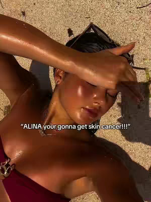 A tan woman lies on a sandy surface, her arm shielding her eyes from the sun. The image is brightly lit, with a focus on her face and upper body. Her skin appears glistening. Text overlayed on the image reads: "ALINA your gonna get skin cancer!!!"