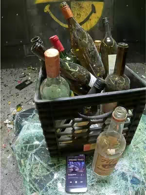 The image shows a black plastic crate filled with empty liquor bottles and a smashed glass surface in the foreground. A cell phone displays a music player application at the bottom. The setup suggests a party or emotional situation.