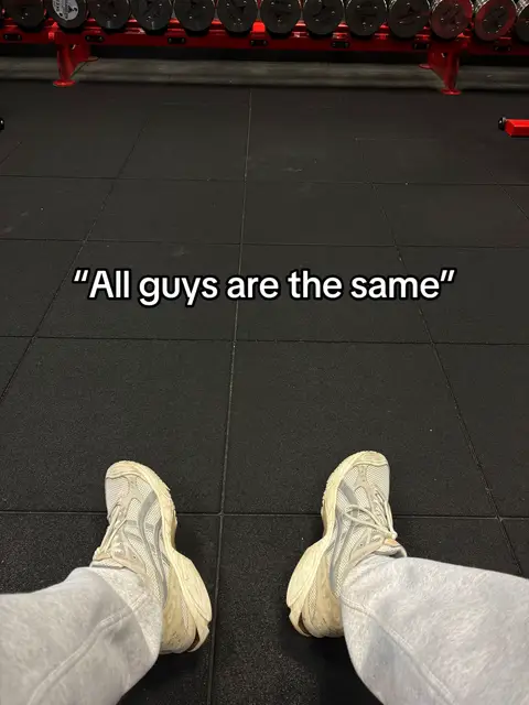 A first-person perspective shot from the floor of a gym. The image shows a person's legs and feet, wearing light-colored sneakers and gray sweatpants. Above, the caption 'All guys are the same' is overlaid in white text, centered on the frame. In the background, a rack of dumbbells in a red frame and a dark floor.