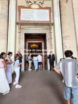 The image shows the entrance to a building with tall columns on either side. The structure has a large stone door and is partly shaded. Several people can be seen in the entrance. There is text overlayed in white.