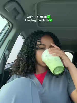 A woman is seated inside a car, holding a green coffee cup up to her mouth. She has curly dark hair and is wearing what appears to be a gray scrubs top and a red shirt underneath. The background is the interior of the car, with a portion of the windshield and the car's roof visible. Text overlays state 'work at 6:30am' and 'time to get matcha'.