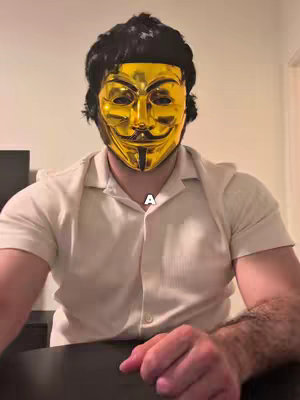 A man wearing a gold Guy Fawkes mask is centered in the frame. He wears a cream-colored button-down shirt and his hands are clasped together. The background is a plain beige wall.