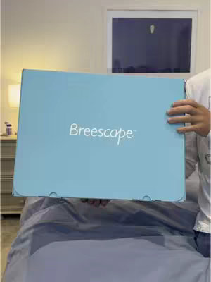 A person holds a light-blue rectangular box, titled with the brand "Breescape" in white script. The box is held on a bed covered with grey bedding. In the background, there is a bedroom setting with a door and a lamp.