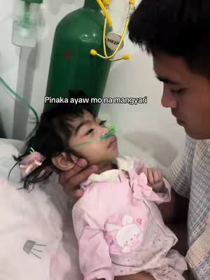 A man is holding a young child who is lying in a hospital bed. They are in a softly lit setting, with a green oxygen tank visible in the background and medical equipment attached to the child. The man's face shows concern as he looks at the child. The child has a tube in their nose and is gazing upwards.