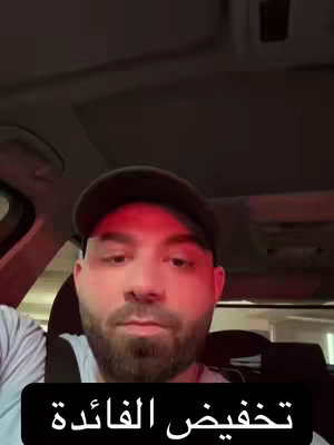 A selfie-style video cover featuring a man inside a car. The man is wearing a dark cap, looking directly at the camera with a serious expression. The lighting is red, casting shadows and a slightly ominous feel. Text in Arabic is added in a black box at the bottom.