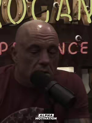 The image is a medium shot featuring a bald man speaking into a microphone. He is wearing a maroon shirt. Behind him, a sign spells out "ROGAN" in a glowing yellow font, and the word "NCE" is visible in red lit-up letters. Below the man, the logo of "ALPHA MOTIVATION" appears in a white font. The lighting suggests an indoor setting.