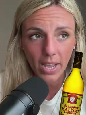 A close-up shot of a woman talking into a microphone, with a bottle of yellow liqueur in the background. Her blonde hair is pulled back, and she appears to be talking and making an expression of shock or disbelief. The bottle, with a prominent label, is positioned to the right of her face.