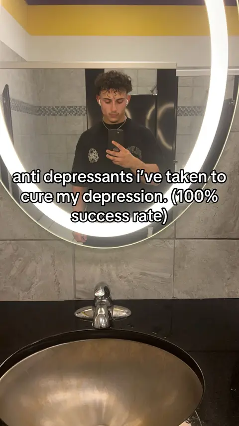 A man is filming himself in a bathroom using his phone. The mirror is a large, illuminated oval, with the man centered and holding the phone up to his face. The background is a clean bathroom setting with a dark counter and a silver faucet below the mirror.