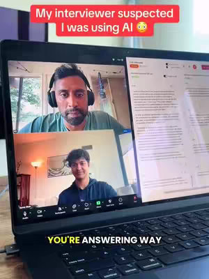 The image captures a laptop screen displaying a Zoom call between three individuals. In the upper portion, the interviewer is visible with headphones. Below, a younger man is present in a casual setting. On the right side, a document is visible, likely the context of the interview. The laptop sits on a work surface with a charging cable and a black desk mat within a well-lit area.