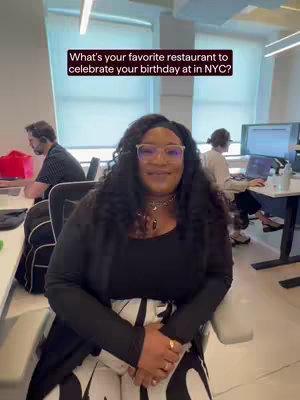 A woman with curly black hair and glasses is sitting in an office setting. She is smiling at the camera. The background includes office desks, windows with natural light, and two people working on computers.