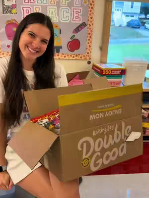 A young woman with long dark hair smiles and sits in a classroom. She is holding a large cardboard box overflowing with popcorn and snacks. The box has text on the side.