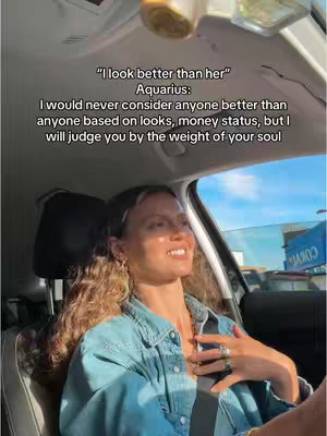 A woman with wavy brown hair is sitting in the front seat of a car, facing slightly towards the right. She is wearing a denim jacket and a light gray top. Text overlays are visible in the image, along with part of the car's interior and an outside view of a building under a blue sky.