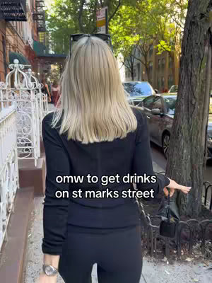 A street scene with the back of a person walking on a sidewalk. The person is blonde and wearing all black with a small black purse. Text overlayed on the image indicates they are going to get drinks on St. Marks Street.